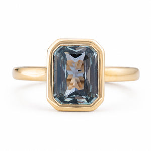 An Antique-inspired, yellow gold solitaire engagement ring featuring a large, faceted emerald-cut blue-gray sapphire in an octagonal bezel setting, shown on a clean white background. 