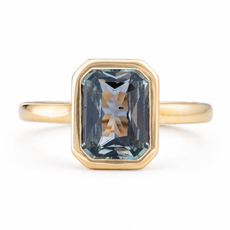 An Antique-inspired, yellow gold solitaire engagement ring featuring a large, faceted emerald-cut blue-gray sapphire in an octagonal bezel setting, shown on a clean white background. 