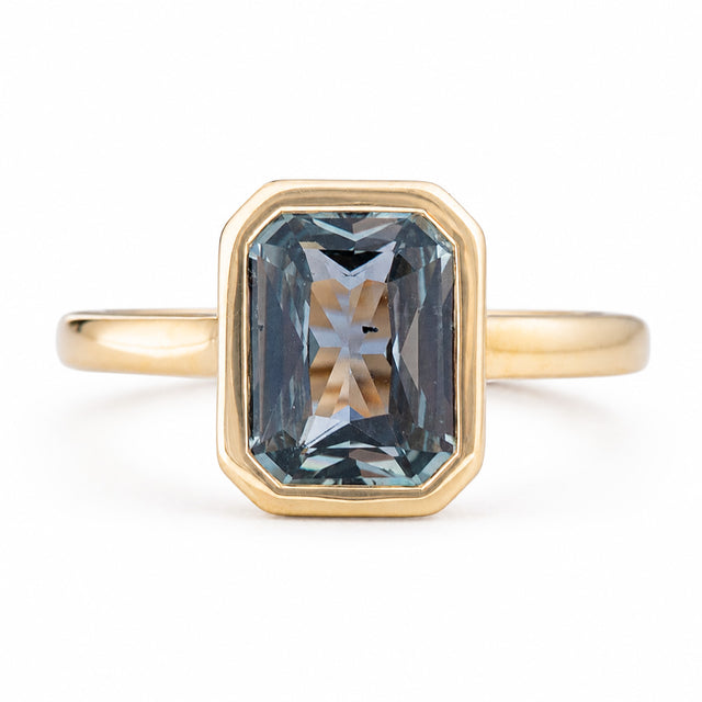 An Antique-inspired, yellow gold solitaire engagement ring featuring a large, faceted emerald-cut blue-gray sapphire in an octagonal bezel setting, shown on a clean white background. 