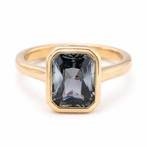 An Antique-inspired yellow gold engagement ring featuring a rectangular bezel setting holding a faceted dark gray-blue teal sapphire, shown on a clean white background.