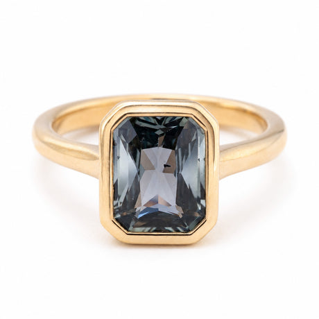 An Antique-inspired yellow gold engagement ring featuring a rectangular bezel setting holding a faceted dark gray-blue teal sapphire, shown on a clean white background.