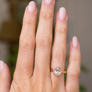 A close-up of a hand wearing an Antique-inspired, delicate gold solitaire engagement ring with a round bezel-set pale pinkish brown sapphire from Sri Lanka.