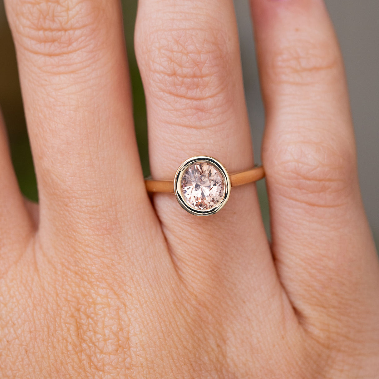 A close-up of a hand wearing a Vintage-inspired yellow gold solitaire ring with a round pale pink sapphire in a bezel setting.