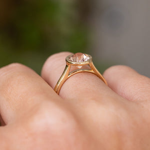 A close-up of a yellow gold solitaire engagement ring worn on a person's finger against a blurred green and gray background.  Displaying the setting and profile of the pale pink round sapphire. 