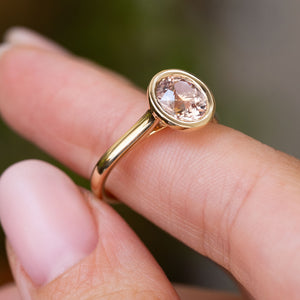 A close-up of an Antique-inspired, round pink sapphire solitaire engagement ring with a yellow gold band and bezel setting, displayed on a finger against a blurred interior background