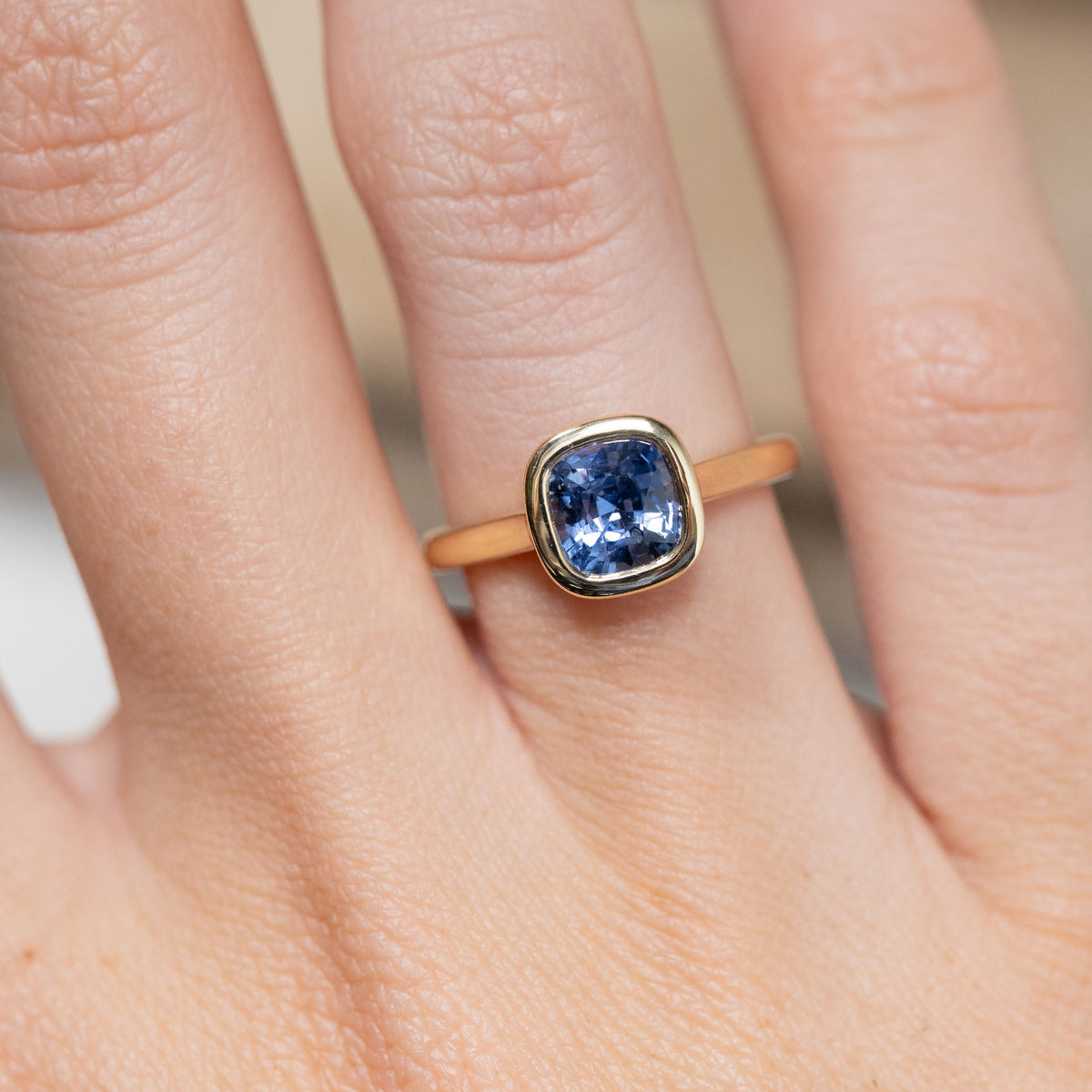 Close-up of the cushion-cut blue sapphire in a smooth yellow gold bezel setting, worn on a finger.