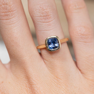 Close-up of the cushion-cut blue sapphire in a smooth yellow gold bezel setting, worn on a finger.
