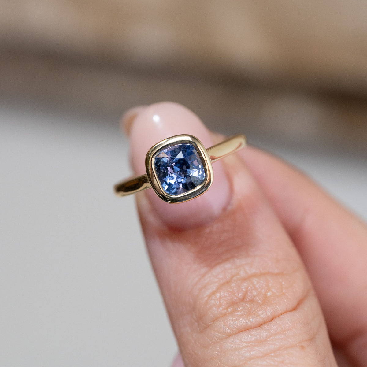 Hand-held close-up of the cushion-cut blue sapphire, framed by a polished yellow gold bezel in an antique-inspired design.