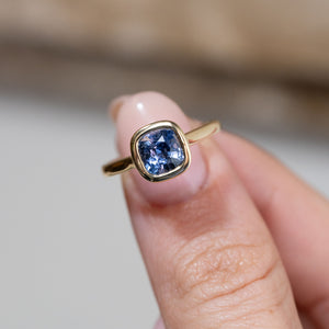 Hand-held close-up of the cushion-cut blue sapphire, framed by a polished yellow gold bezel in an antique-inspired design.