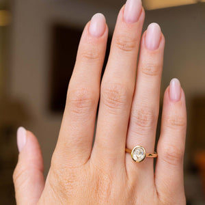 A close-up of a hand wearing a vintage-inspired, pale yellow sapphire solitaire engagement ring in yellow gold, set against a softly blurred indoor background.