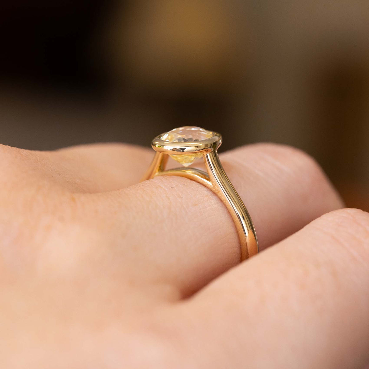 A close up of the side profile of an Antique-inspired, pale yellow sapphire solitaire engagement ring worn on a finger.  Showcasing the yellow gold setting and band which allows a wedding band to sit closely alongside the ring once worn. 