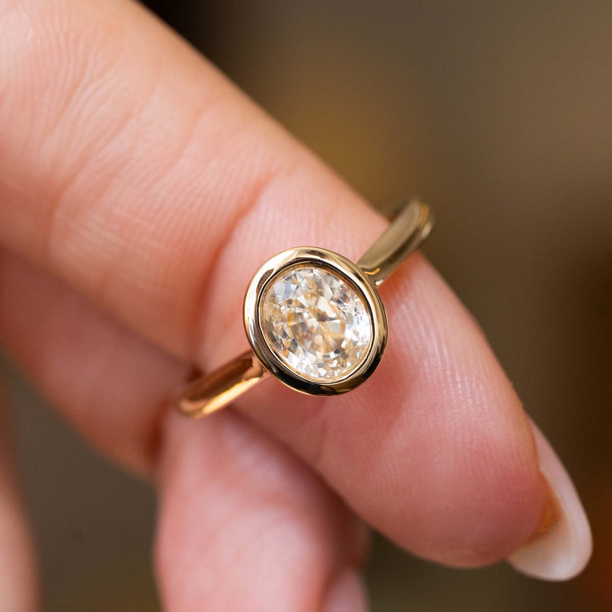 A close-up of a yellow gold antique-inspired solitaire engagement ring with a bezel-set yellow sapphire, worn on a person's finger against a softly blurred warm brown background.