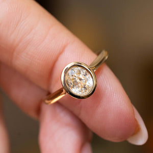 A close-up of a yellow gold antique-inspired solitaire engagement ring with a bezel-set yellow sapphire, worn on a person's finger against a softly blurred warm brown background.