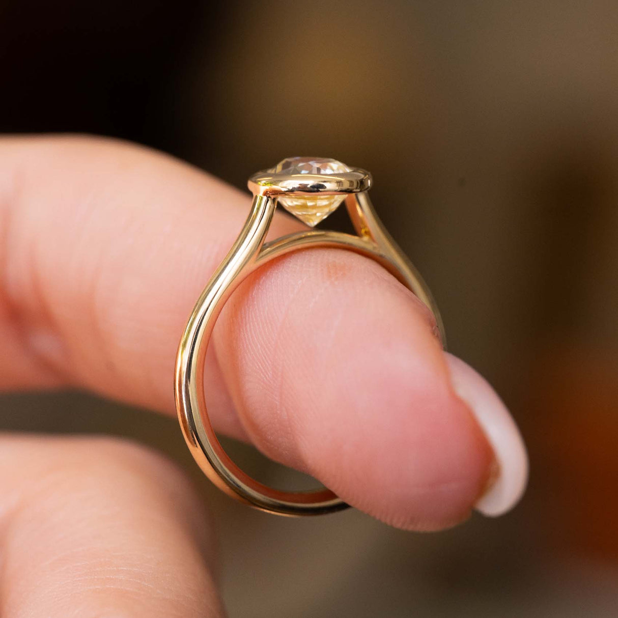 A close-up of a vintage-inspired yellow gold ring set with a yellow sapphire showcasing the yellow gold band and wedding fit design.  Held on a person's finger against a blurred warm brown background.