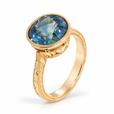 An angled photograph of the Montana sapphire ring, revealing its teal gemstone, decorative gold shoulders and raised setting.