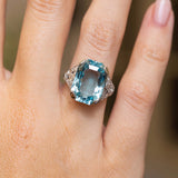 Close-up of an Art Deco platinum ring featuring a faceted light blue aquamarine flanked by intricate round diamond shoulders.
