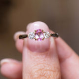 Vintage, 1990s pink sapphire and diamond three-stone engagement ring in platinum held in between the thumb and finger against a blurred background.