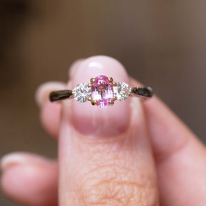 Vintage, 1990s pink sapphire and diamond three-stone engagement ring in platinum held in between the thumb and finger against a blurred background.