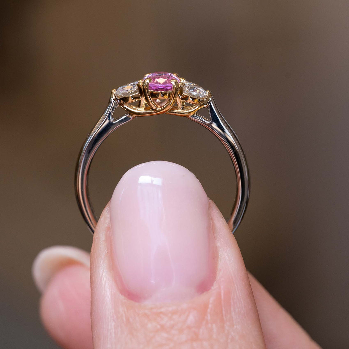 A close up image of a trilogy Vintage 1990's engagement ring in platinum with a yellow gold setting held up to showcase the profile of the central pink sapphire and two diamonds against a blurry background.