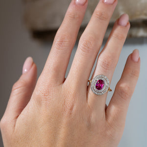 Antique Edwardian Burmese ruby and diamond cluster engagement ring worn on a hand, showcasing its vibrant pinkish-red ruby, double diamond halo and elegant yellow gold band.