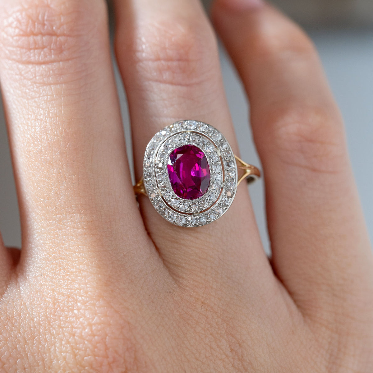 Close-up of an antique Edwardian Burmese ruby and diamond cluster ring worn on a finger, featuring a rich pinkish-red oval ruby framed by two rows of diamonds in platinum.
