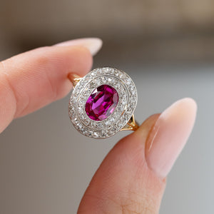 Detailed close-up of an antique Edwardian Burmese ruby and diamond cluster engagement ring held between fingers, showcasing the oval ruby, concentric diamond halos and delicate platinum detailing.