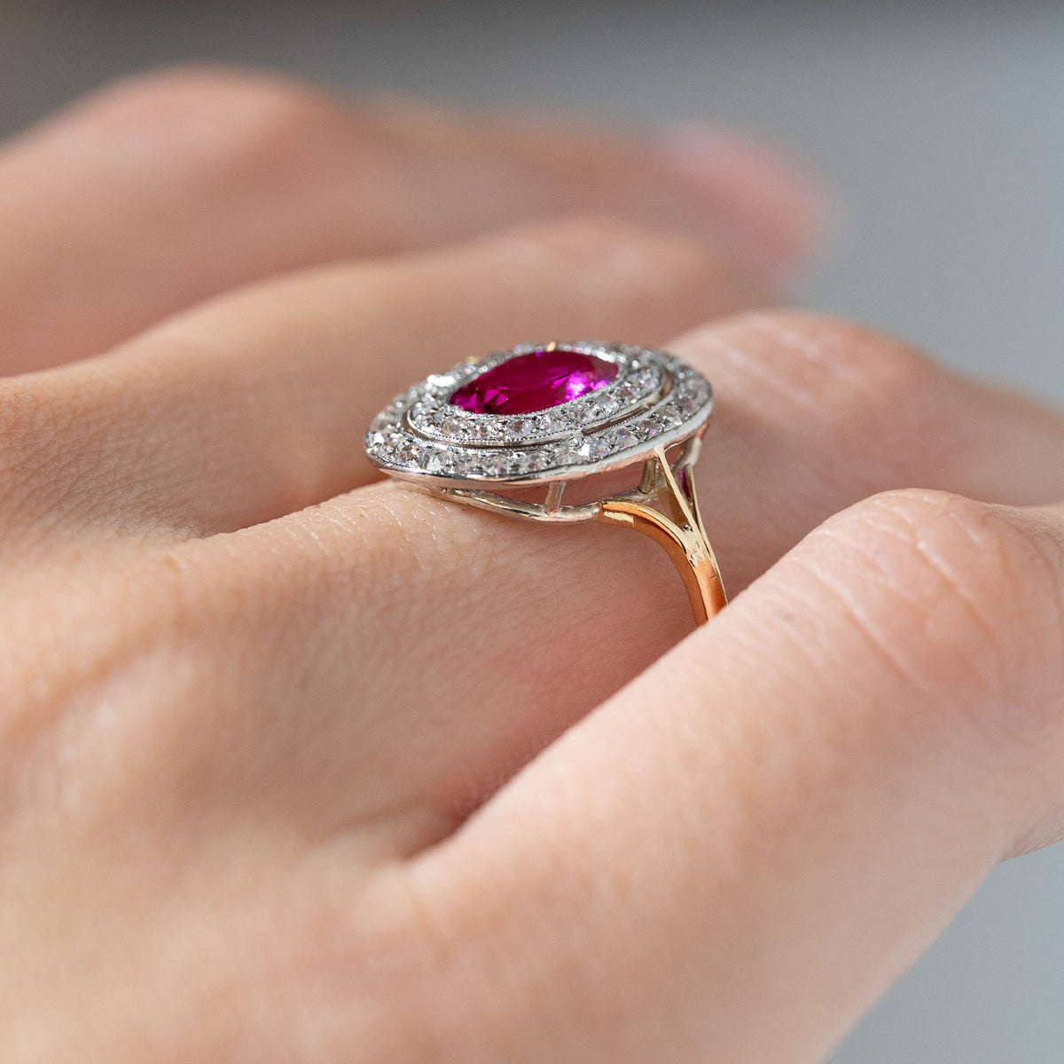 Side angle of an antique Edwardian Burmese ruby and diamond cluster ring worn on a finger, revealing its raised double diamond halo, open gallery and yellow gold band.