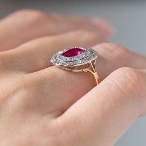 Side angle of an antique Edwardian Burmese ruby and diamond cluster ring worn on a finger, revealing its raised double diamond halo, open gallery and yellow gold band.