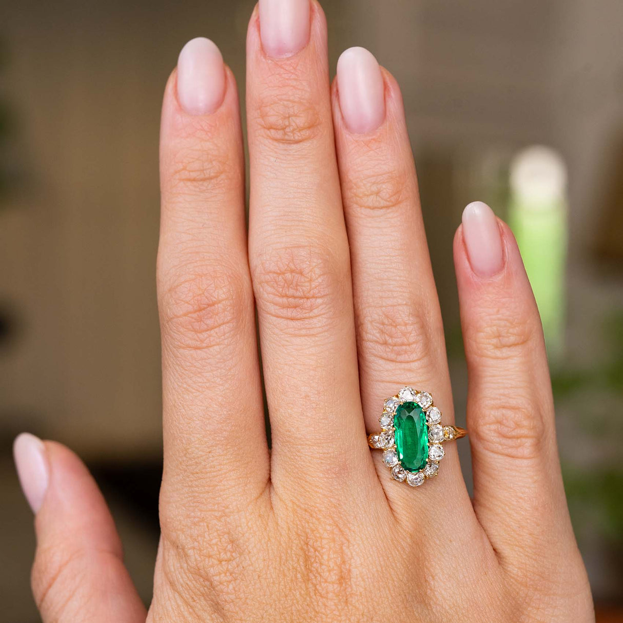 A close-up of a hand wearing an Antique, Belle Époque Colombian emerald and diamond cluster engagement ring with a yellow gold band. 