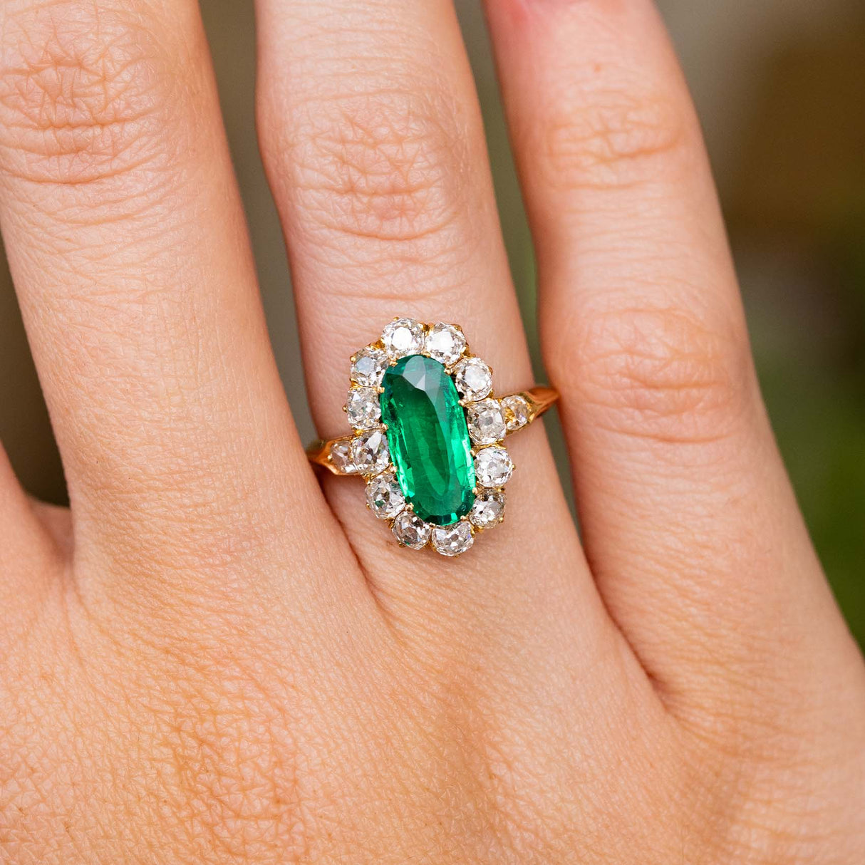 A close-up of an Antique, Belle Époque emerald and diamond cluster engagement ring, worn on a finger and showcasing the Colombian emerald and halo of mine-cut diamonds that surround it.  