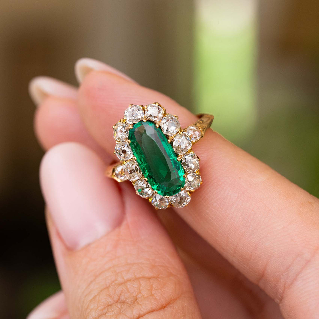 A close-up of a hand holding an Antique, Belle Époque cluster engagement ring, set with a large elongated oval Colombian emerald, surrounded by a halo of diamonds with a yellow gold band, set against a softly blurred green background