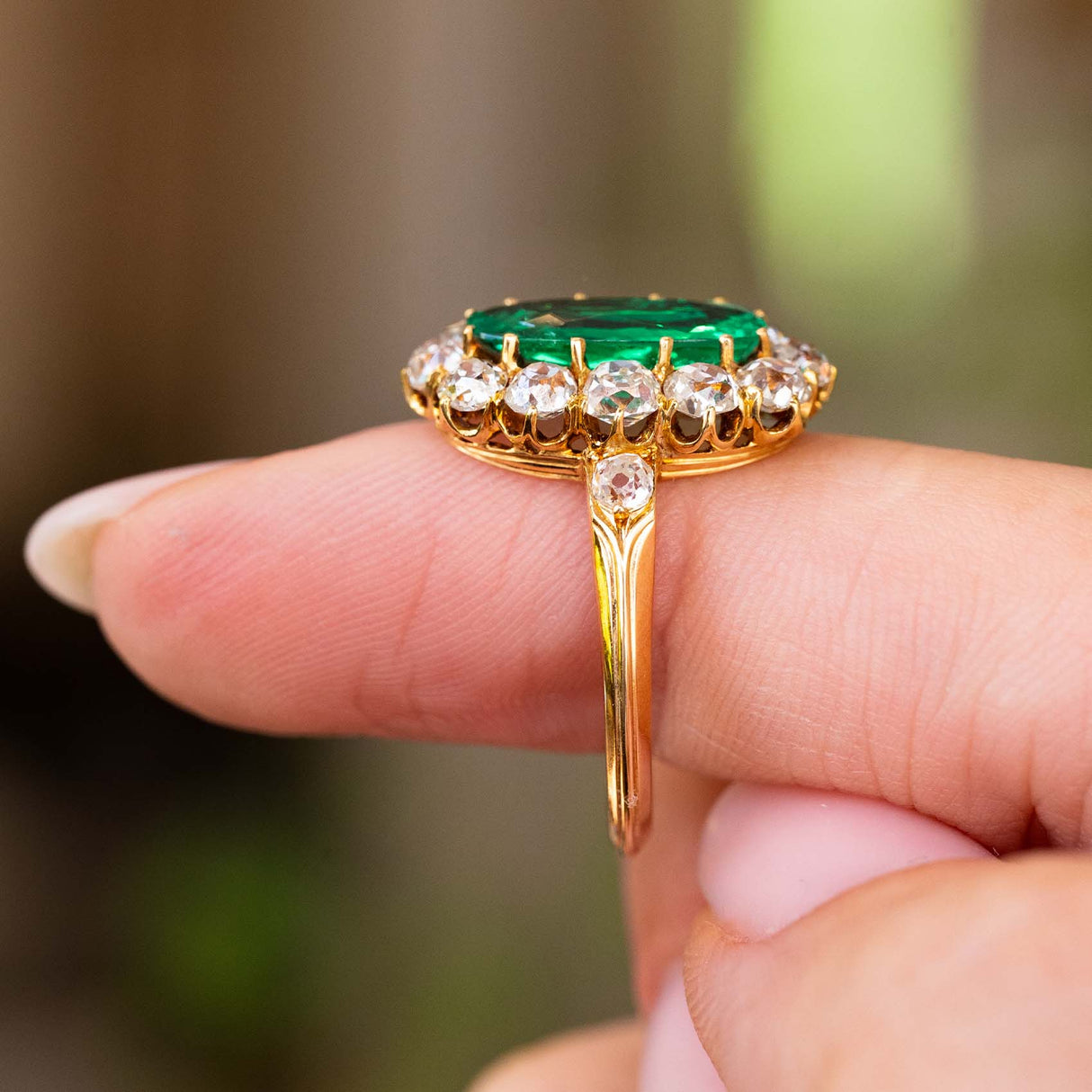 A close up side profile of an Antique Belle Époque yellow gold cluster engagement ring.  Showing the setting of the Colombian emerald and diamonds and the band.  Held on a finger against a blurred interiors background. 