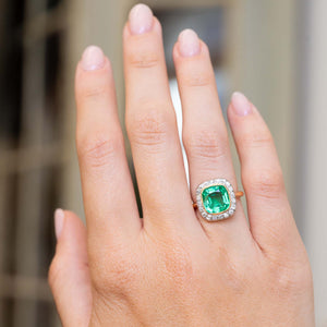 An image of a hand outstretched wearing an antique Belle Époque era yellow gold engagement ring featuring an emerald and diamonds worn on the finger against a blurred background.