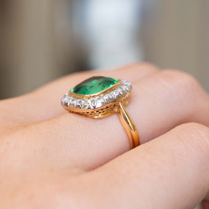 A zoomed in image of an antique Belle Époque era emerald and diamond coronet cluster yellow gold engagement ring worn on the finger and shown at an angle to display how the ring sits on the finger when worn. 