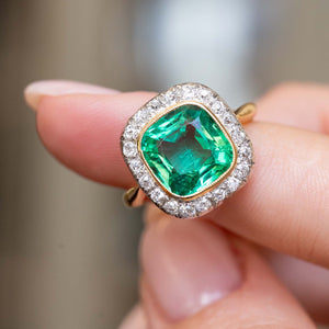 A close-up image of an antique green emerald and diamond cluster engagement ring in yellow gold resting on a finger against a blurry background. 