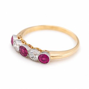 A three-quarter close-up of an Antique, Edwardian pink-ruby and old-cut diamond five-stone ring with a platinum setting and a yellow gold band with a pierced gallery.