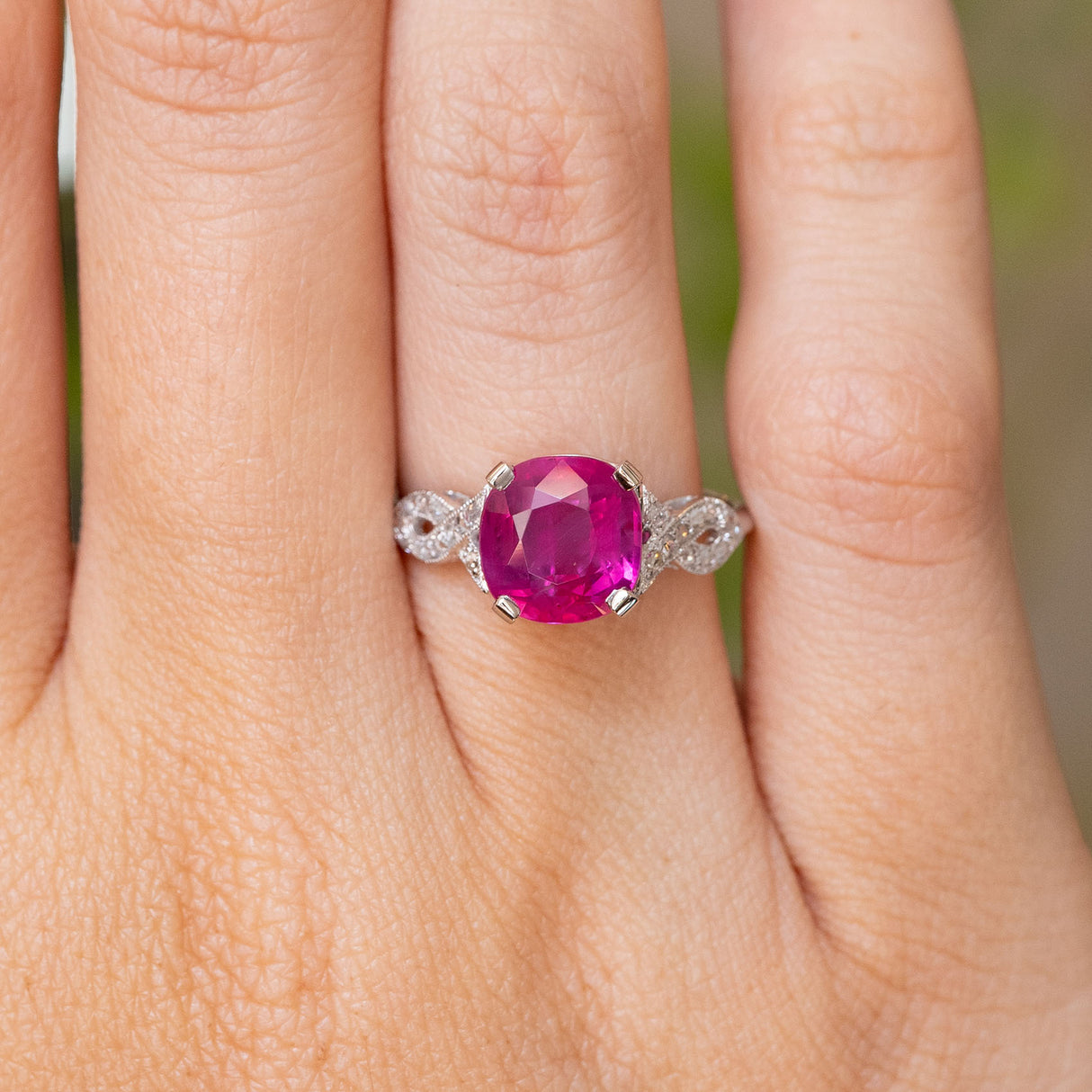 A close-up of a hand wearing an Antique Art Deco white gold engagement ring with a large, cushion-cut, vibrant pink central ruby and small diamond stones on the shoulders.