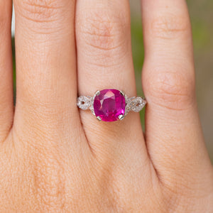 A close-up of a hand wearing an Antique Art Deco white gold engagement ring with a large, cushion-cut, vibrant pink central ruby and small diamond stones on the shoulders.