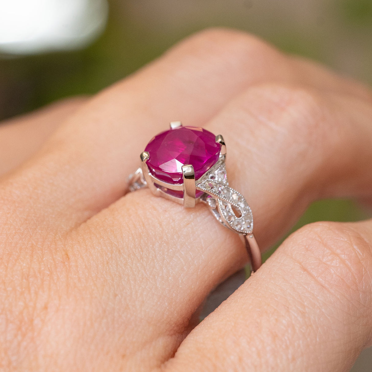 A close-up of a hand wearing an Antique Art Deco engagement ring with a large cushion-cutl pink ruby in a white-gold setting with diamonds set  along the band.