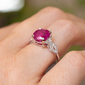 A close-up of a hand wearing an Antique Art Deco engagement ring with a large cushion-cutl pink ruby in a white-gold setting with diamonds set  along the band.