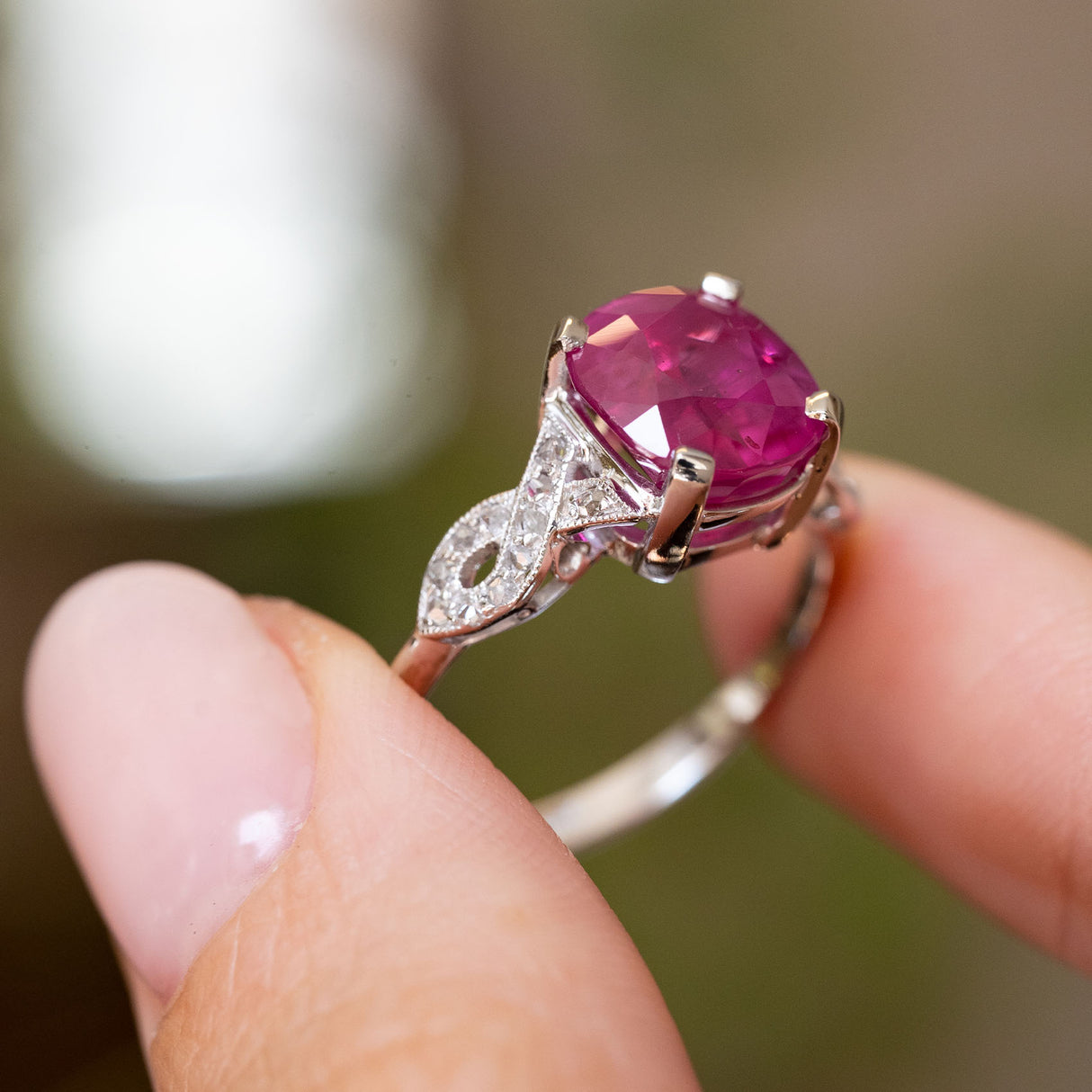 A close-up of an Antique Art Deco flanked solitaire engagement ring.  Showcasing the central pink ruby and diamond accents on the band, held between two fingers against a blurred background. 