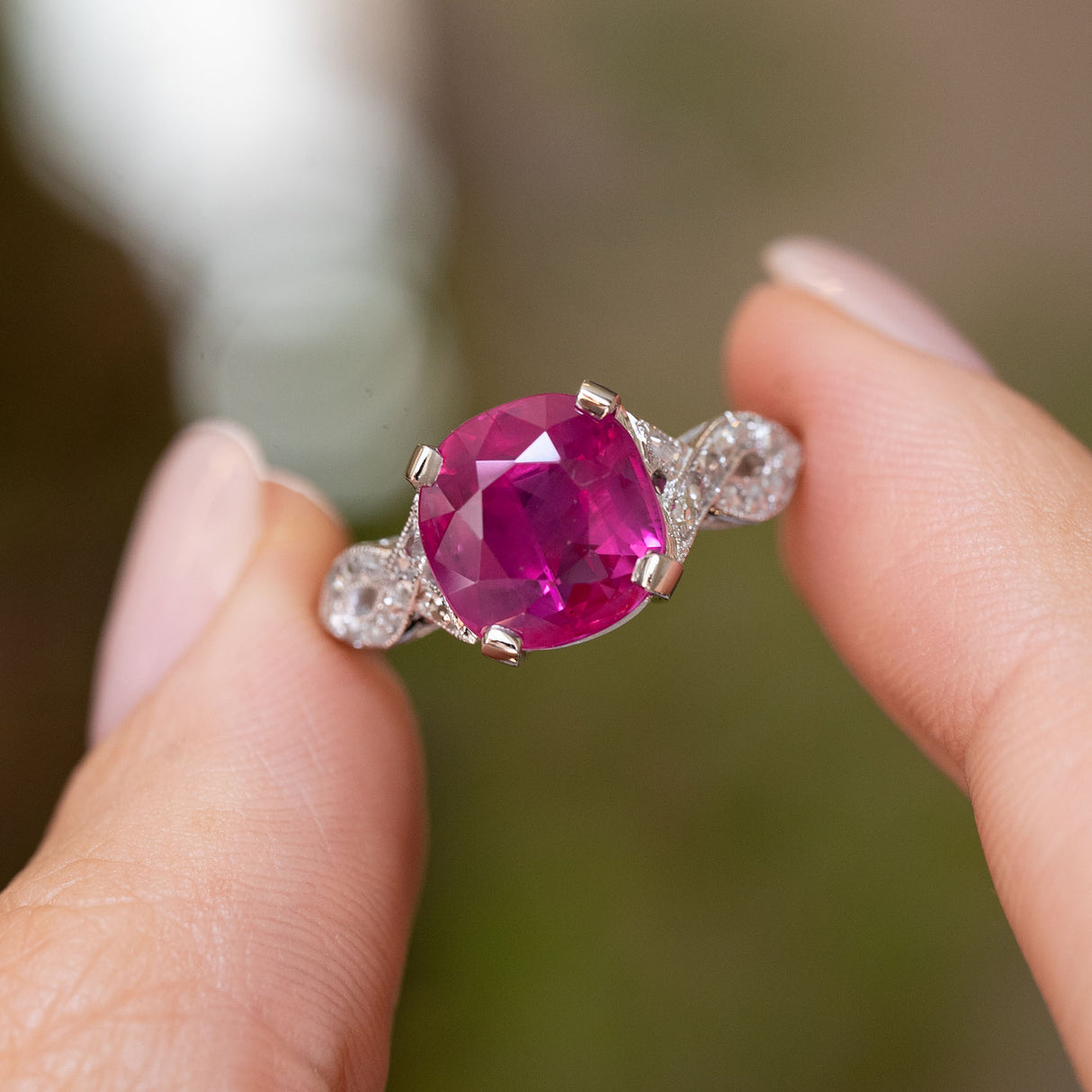 A close-up shot of a flanked solitaire engagement ring held between two fingers. The Antique Art Deco ring features a large Burmese pink ruby flanked by smaller diamond accent stones on a blurred green and brown background.