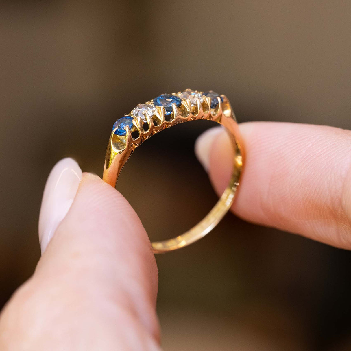 Gallery detail of an antique Edwardian sapphire and diamond five-stone ring held between the fingers.