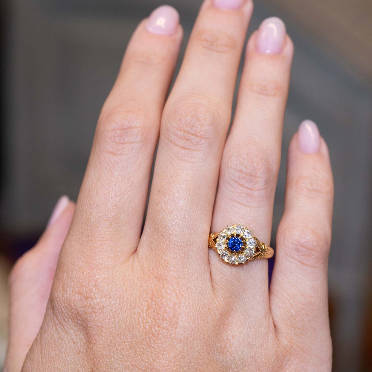 A picture of an Antique, Victorian blue sapphire and old mine-cut diamond cluster engagement ring in yellow gold worn on the finger and shown on an outstretched hand to show the proportions.