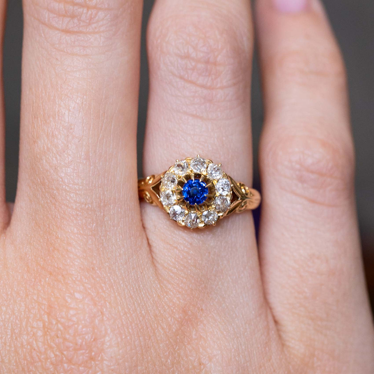 A close-up of an Antique, Victorian blue sapphire and old mine-cut diamond cluster ring in yellow gold worn on the finger and shown close up.