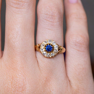 A close-up of an Antique, Victorian blue sapphire and old mine-cut diamond cluster ring in yellow gold worn on the finger and shown close up.