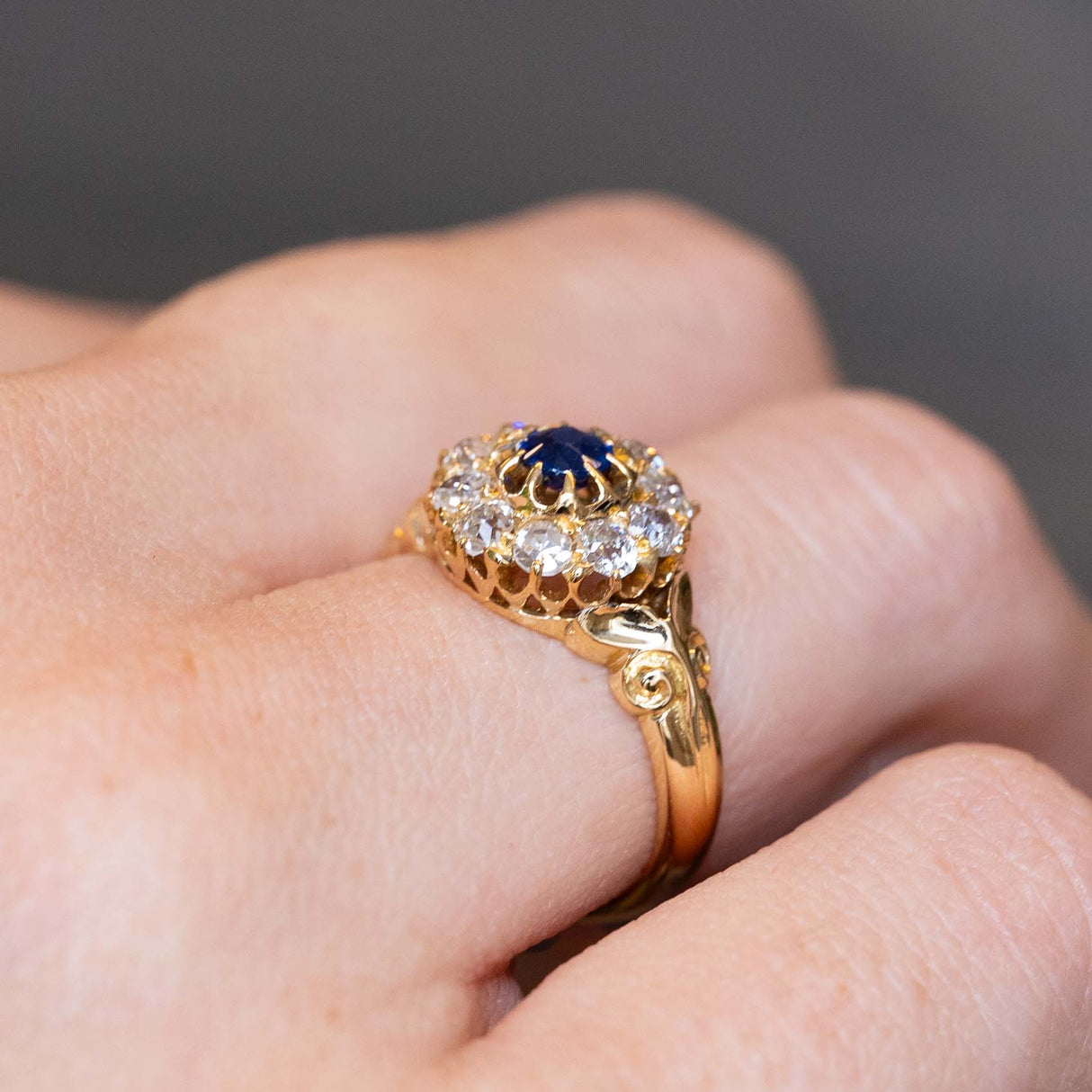 An Antique Victorian blue sapphire and old mine-cut diamond cluster engagement ring in yellow gold worn on the finger and seen at an angle to show off the decorative details and setting style, worn on the finger and shown up close.