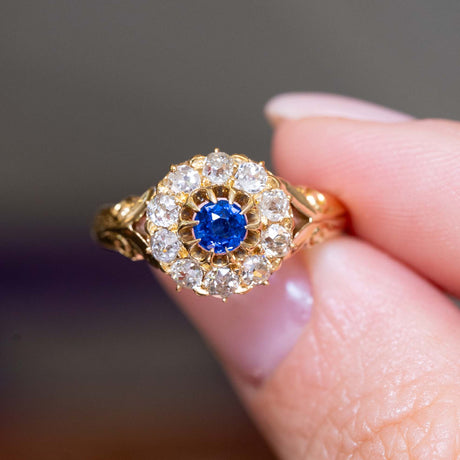 A close-up of an Antique, Victorian blue sapphire and old cut diamond cluster engagement ring held between the fingers against a blurred gray background.