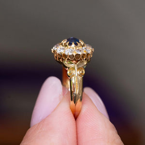 A close-up image of an Antique, Victorian blue sapphire and diamond cluster engagement ring in yellow gold ring held between fingers and turned to the side to showcase the gold scrolling decorative shoulders and setting style.