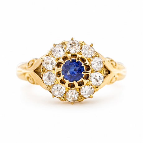 An Antique, Victorian sapphire and old cut diamond cluster ring in yellow gold featuring decorative shoulders shown against a plain white background.
